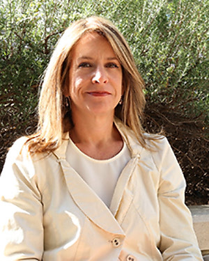 A blonde woman sitting on a bench wearing a cream-colored blazer.